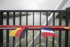 Eine Aufnahme von der abwärts gelegenen Seite des Damms, mit im Vordergrund dem Geländer der Fußgängerbrücke mit französischen und deutschen Flaggen und im Hintergrund der Landschaft des Oberen Rheins.