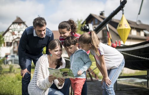 Eine Familie macht die Schatzsuche in Rheinau. Die Mutter hält das Heft in den Händen, während die drei Kinder und der Vater aufmerksam lesen und zuhören.